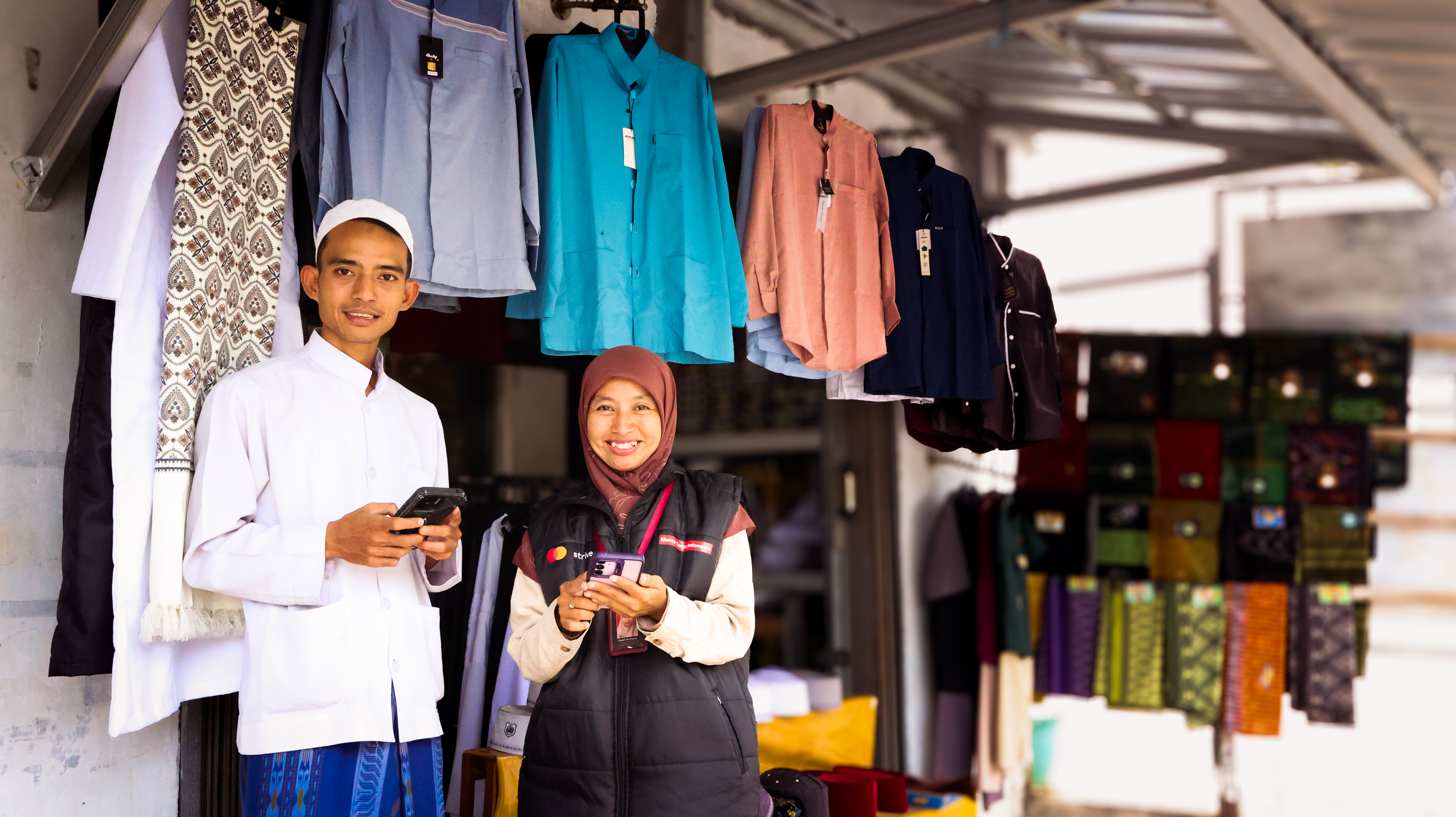 Two indonesians stand together inside clothing shop holding phones and smiling.