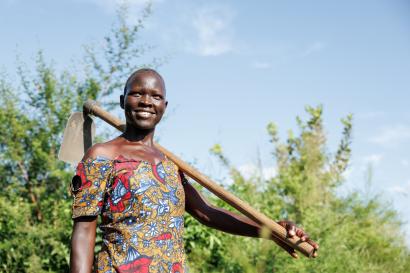 Ugandan woman stands in field smiling with farm implement over her shoulder.