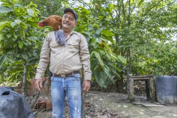 Colombia man stands amidst agricultural setting with rooster on his shoulder.