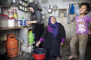 An adult stirs a pot while smiling at a happy child while another adult preps ingredients nearby.