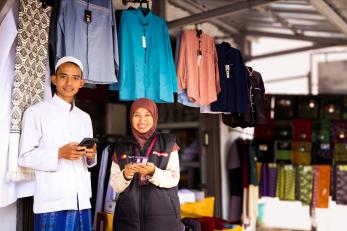 Two Indonesians stand together inside clothing shop holding phones and smiling.