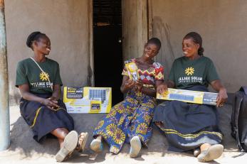 Ugandan women on front porch of home with new, boxed solar panel systems.