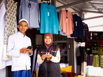 Two indonesians stand together inside clothing shop holding phones and smiling.
